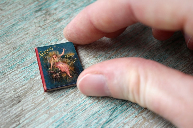 May include: A miniature hardcover book titled "Grimms Fairy Tales" with a red spine and a blue cover featuring an illustration of a fairy with wings and a wand. The fairy is wearing a pink dress and is sitting on a branch.