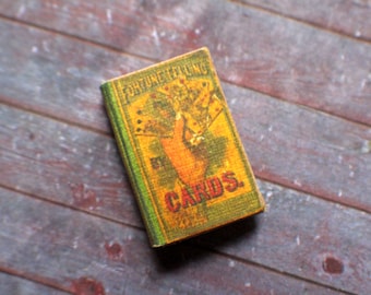 May include: A miniature book titled "Fortune Telling by Cards" with a green spine and a yellow cover featuring a hand holding cards. The book is set against a wooden surface with a rustic appearance.