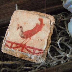 May include: A rectangular, aged-looking piece of art featuring a red peacock illustration. The bird stands on a red fence against a beige background. The artwork is set in a wooden box with dried plant material.