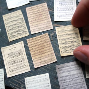 May include: Miniature sheet music pages with aged paper appearance. The pages are arranged on a dark surface, with one being held by a finger. The music notes are printed in black ink, creating a vintage aesthetic. Ideal for crafting or scrapbooking.