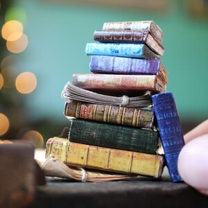 May include: A stack of miniature books with colorful covers. The books are in various colors, including blue, brown, green, and yellow. Some books have aged, textured covers, and others have smooth, solid colors. A hand is holding one of the books.
