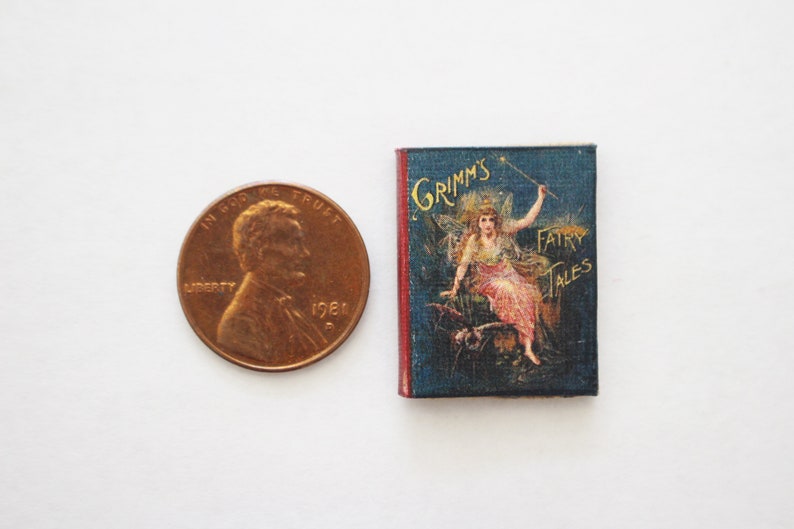 May include: A miniature book with a blue cover and gold lettering. The cover features an illustration of a fairy with a wand, sitting on a cloud. The title of the book is "Grimm's Fairy Tales".