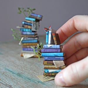 Op de afbeelding: Miniatuur stapels boeken met blauwe, bruine en paarse covers. Een kleine vlinder rust bovenop een stapel. Groene takjes zijn verspreid tussen de boeken. De boeken zijn gerangschikt op een houten oppervlak.