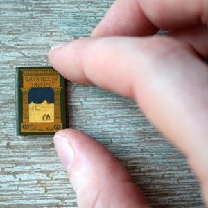 May include: Miniature book titled "The Spell of Egypt" with a green cover and gold lettering. The cover features an illustration of a desert scene with pyramids and camels. The book is held between two fingers.