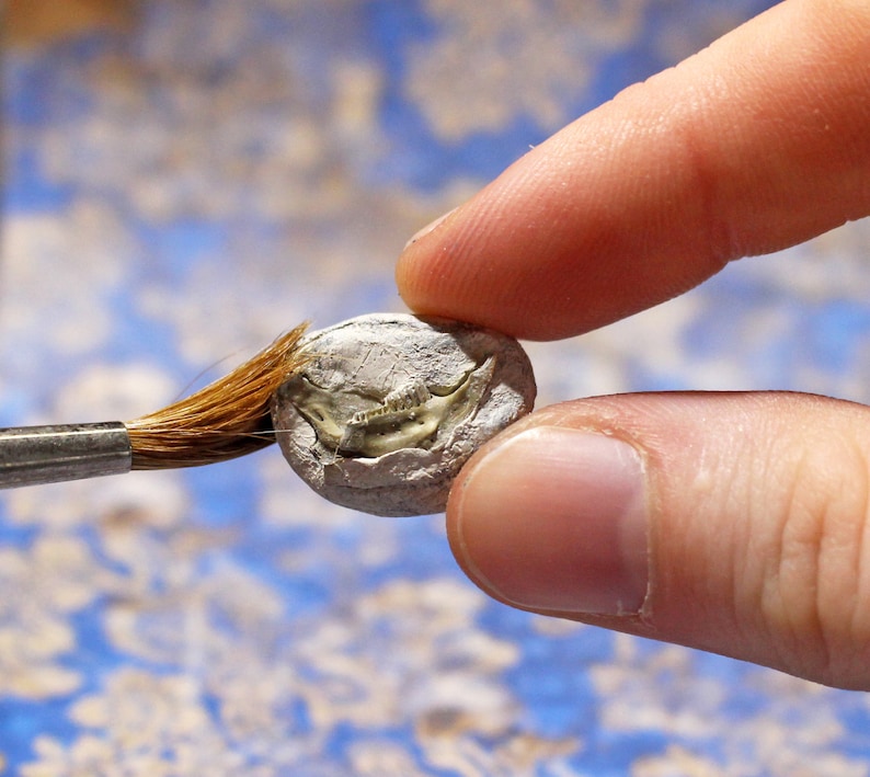 May include: A small, round, gray fossil being brushed with a fine-tipped brush. The fossil appears to be of a small animal, with visible bone structure. The background is a blurred pattern of blue and white.