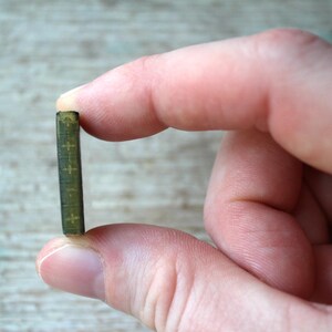 May include: A miniature, olive green book with gold cross details held between fingers. The tiny book has a textured cover and appears to be a religious item or a piece of dollhouse decor. The background is a light wood surface.