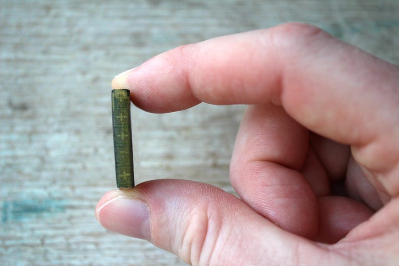 May include: A miniature, olive green book with gold cross details held between fingers. The tiny book has a textured cover and appears to be a religious item or a piece of dollhouse decor. The background is a light wood surface.