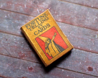 May include: A miniature book titled "Fortune Telling by Cards" with a weathered, vintage appearance. The cover is tan with black text and a red and black illustration of a wizard. The book is set against a wooden background.