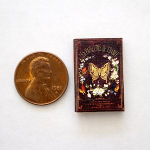 May include: A miniature book with a burgundy cover, featuring the title "LES PAPILLONS DE FRANCE" and a gold butterfly illustration. The book is next to a U.S. penny for size comparison.