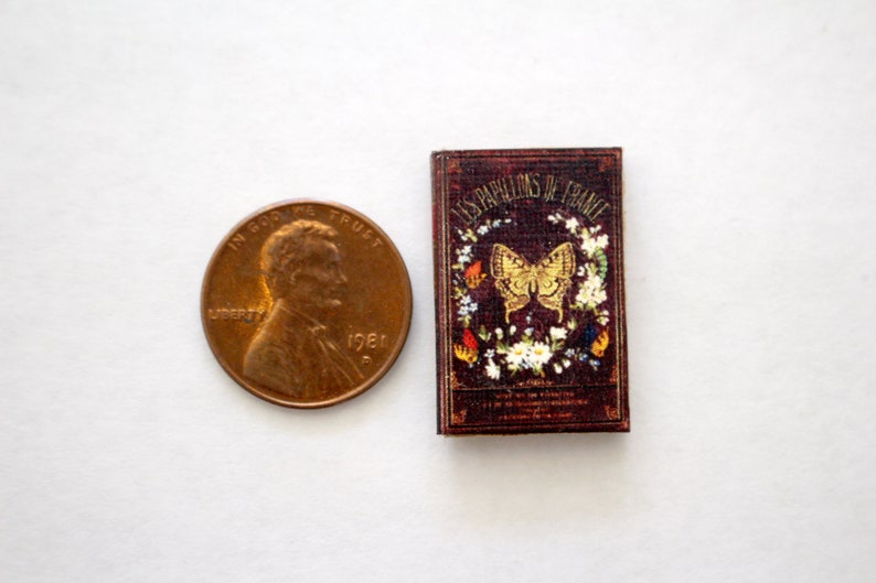 May include: A miniature book with a burgundy cover, featuring the title "LES PAPILLONS DE FRANCE" and a gold butterfly illustration. The book is next to a U.S. penny for size comparison.