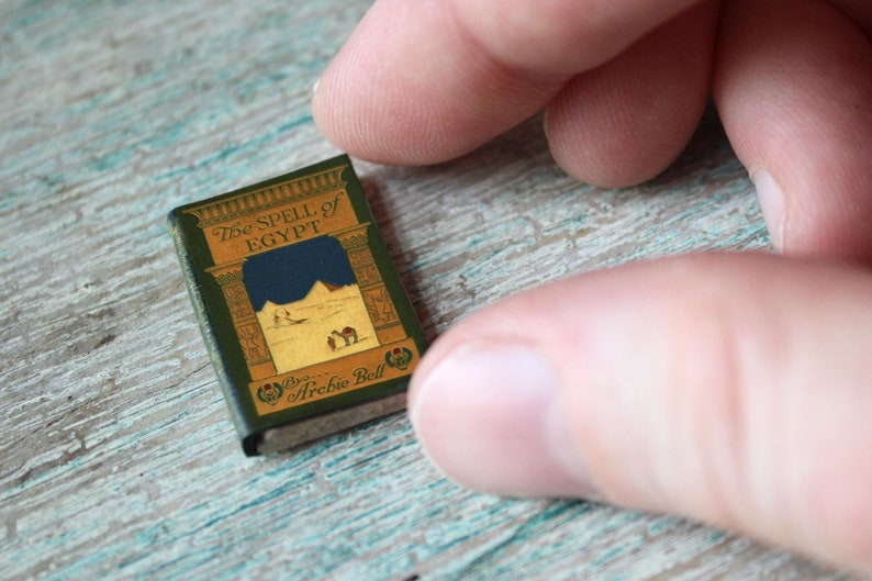 May include: A miniature book titled "The Spell of Egypt" with a green cover and gold lettering. The cover illustration depicts pyramids and a desert scene. The book is held between two fingers.