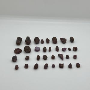 May include: A collection of small, rough, red and purple gemstones. The gemstones are various shapes and sizes, and some have a slightly metallic sheen.