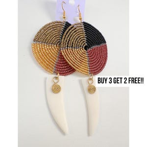 May include: Handmade beaded earrings with a circular design in gold, black, and red. The earrings have a white, tooth-shaped pendant and gold-colored hooks. The text "BUY 3 GET 2 FREE!!" is visible.