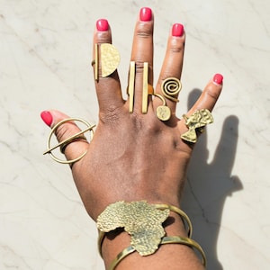 May include: A hand displaying various gold-toned rings and a bracelet. The rings feature geometric shapes, spirals, and a bust. The bracelet has an African continent design. The nails are painted a bright red.