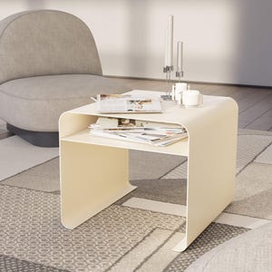 Puede incluir: Una mesa auxiliar moderna de color crema con un diseño curvo. La mesa tiene un estante inferior y contiene revistas y algunos objetos decorativos. Un sillón gris está en el fondo, junto con una alfombra estampada.