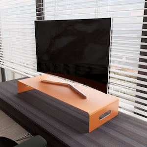 May include: An orange-colored monitor stand with a curved design and cut-out handles. A large black television screen sits on top of the stand. The stand is placed on a dark wood-grain surface near a window with white blinds.