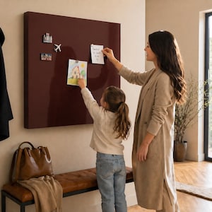 May include: A large, rectangular, burgundy-colored magnetic board mounted on a wall. Two people are placing notes and drawings on the board. Decorative magnets are attached. A bench and a leather bag are in the foreground.