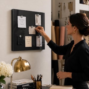 May include: A black magnetic board with silver magnets holding notes and fabric swatches. The notes include text: "Design Review 10 AM Studio B" and "Client Fitting Thursday 4 PM". A woman in a black shirt is reaching towards the board.