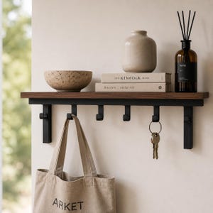 May include: A wall-mounted shelf with a dark wood top and a black metal frame featuring six hooks. On the shelf sits a textured bowl, a beige vase, two books titled The Kinfolk Home and Interiors Atelier AM, and a brown glass diffuser bottle.