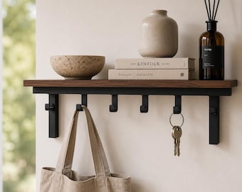Könnte beinhalten: Ein Wandregal mit einer dunklen Holzplatte und einem schwarzen Metallrahmen mit sechs Haken. Auf dem Regal stehen eine strukturierte Schale, eine beige Vase, zwei Bücher mit den Titeln The Kinfolk Home und Interiors Atelier AM sowie ein Diffusor.