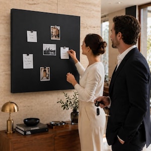 May include: A large, square black magnetic board with several small notes and photos attached. A woman and a man are standing in front of the board, with the woman appearing to be adjusting a note. The board is mounted on a light-colored wall.