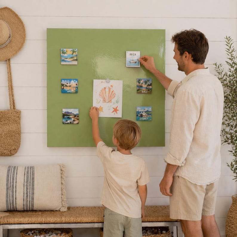 May include: A green magnetic board displays travel-themed magnets and a seashell drawing. A man and a child are arranging the magnets, which feature scenic views and the names of locations like Maui, Ibiza, and Sydney. A straw hat and tote bag hang on the wall.