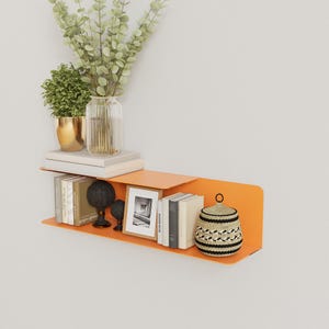 May include: An orange metal floating shelf with books, decorative objects, and a woven basket. The shelf holds a framed picture, a black sphere, and a small black decorative object. A vase with greenery and a potted plant sit above.