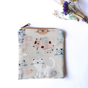 Op de afbeelding: Een beige etui met ritssluiting met een herhaald patroon van cartoonkatten in verschillende kleuren. De katten worden afgebeeld met verschillende uitdrukkingen en poses, sommige met bollen wol. De rits is bruin.