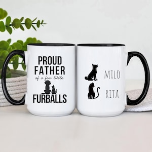 May include: Two white ceramic mugs with black handles and black rims. The mug on the left says "Proud Father of a few little Furballs" with a silhouette of a dog and cat. The mug on the right says "Milo" and "Rita" with silhouettes of a dog and cat.