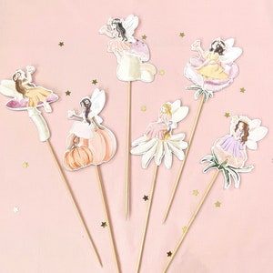 May include: Six colorful fairy cake toppers with sticks. Each topper features a fairy with wings and a flower, pumpkin, or mushroom design. The fairies are wearing dresses in shades of pink, yellow, and purple.