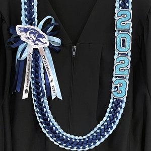 May include: A graduation lei in navy blue, light blue, and white, with the year "2023" in glittery light blue numbers. The lei features ribbons with text and a falcon emblem, worn over a black graduation gown.