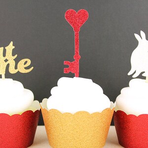 May include: Three cupcakes decorated with different toppers. The cupcake on the left has a gold glitter "eat me" topper. The cupcake in the middle has a red glitter heart-shaped key topper. The cupcake on the right has a white glitter rabbit topper.