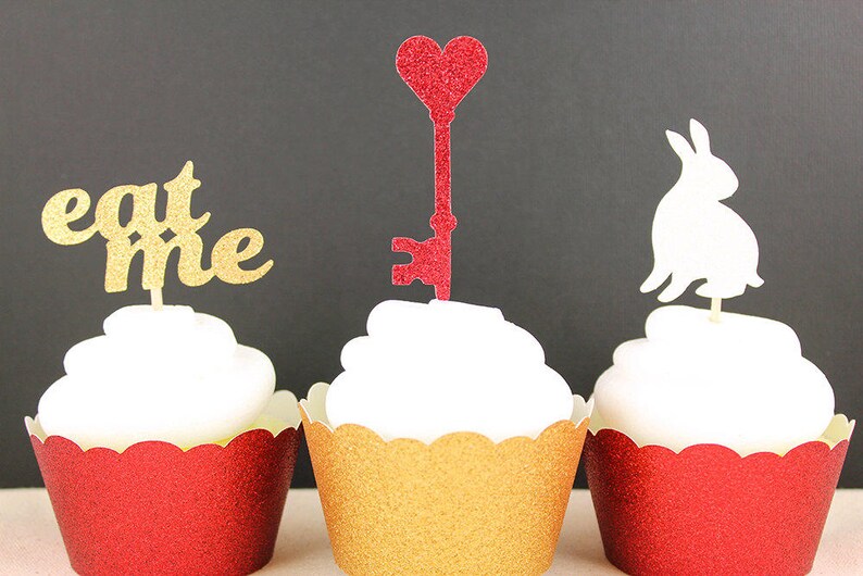 May include: Three cupcakes decorated with different toppers. The cupcake on the left has a gold glitter "eat me" topper. The cupcake in the middle has a red glitter heart-shaped key topper. The cupcake on the right has a white glitter rabbit topper.