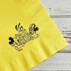 May include: Yellow paper napkin featuring a black and white illustration of Mickey Mouse, Minnie Mouse, and Pluto playing with alphabet blocks. The text "&copy;Disney" is printed below the illustration.