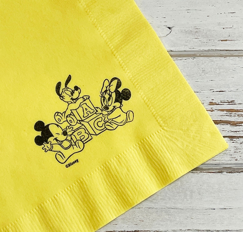 May include: Yellow paper napkin featuring a black and white illustration of Mickey Mouse, Minnie Mouse, and Pluto playing with alphabet blocks. The text "&copy;Disney" is printed below the illustration.