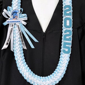 May include: A graduation lei in light blue and white, with the year "2025" in glittery blue letters. A bow with the school's logo and the words "Warriors" and "Congratulations Micah!" are also visible.