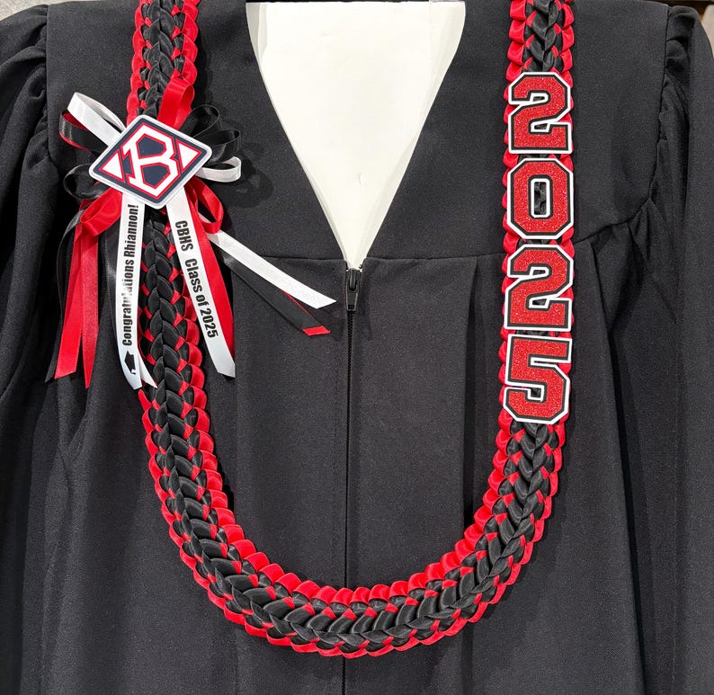 May include: A black and red graduation lei with the text "2025" in red and white. The lei is braided with black and red ribbons. A white ribbon with the text "Congratulations Rhiannon!" and "CBHS Class of 2025" is attached.