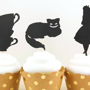 May include: Three cupcakes decorated with black silhouette toppers. The toppers are of a teacup, a Cheshire cat, and Alice from Alice in Wonderland. The cupcakes are in brown cupcake liners with gold polka dots.