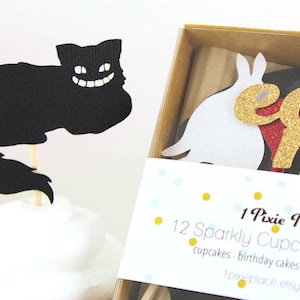 May include: A set of 12 sparkly cupcake toppers featuring a black Cheshire Cat, a white rabbit, and gold glitter letters spelling "eat me". The toppers are packaged in a brown box with a white label that reads "1 Pixie Place 12 Sparkly Cupcake Toppers cupcakes - birthday cakes - appetizer picks 1pixieplace.etsy.com"