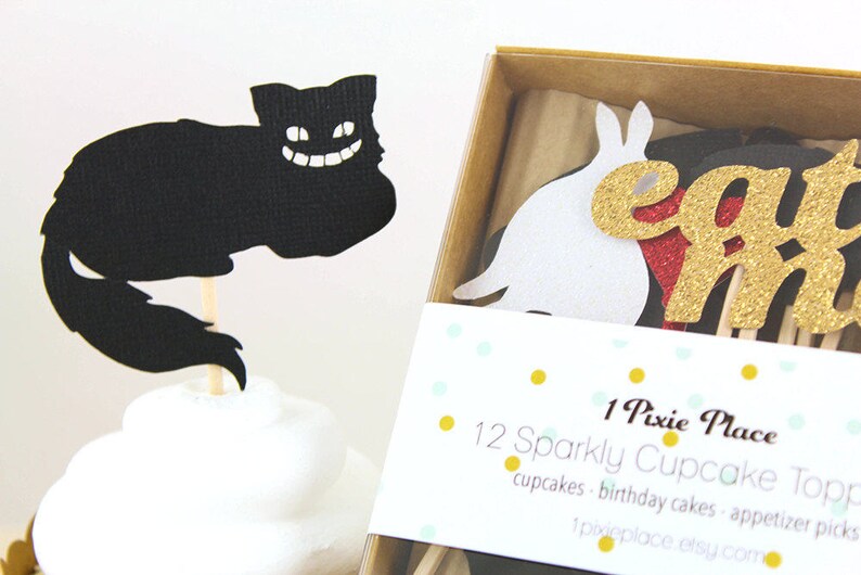 May include: A set of 12 sparkly cupcake toppers featuring a black Cheshire Cat, a white rabbit, and gold glitter letters spelling "eat me". The toppers are packaged in a brown box with a white label that reads "1 Pixie Place 12 Sparkly Cupcake Toppers cupcakes - birthday cakes - appetizer picks 1pixieplace.etsy.com"