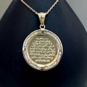 May include: A gold-colored pendant necklace with a circular charm. The charm features an inscription surrounded by small, clear stones. The necklace is displayed on a black, teardrop-shaped stand.