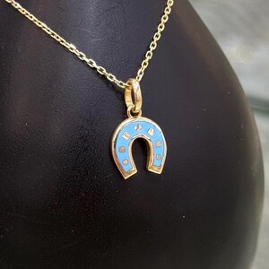 Puede incluir: Un collar de cadena dorada con un colgante de herradura con incrustaciones de esmalte azul claro y pequeños puntos dorados. El colgante cuelga de una delicada cadena de eslabones dorados, mostrando un acabado metálico pulido y un contraste azul.
