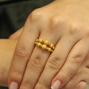 May include: A gold ring with a textured design featuring multiple small round beads. The ring is worn on a finger.