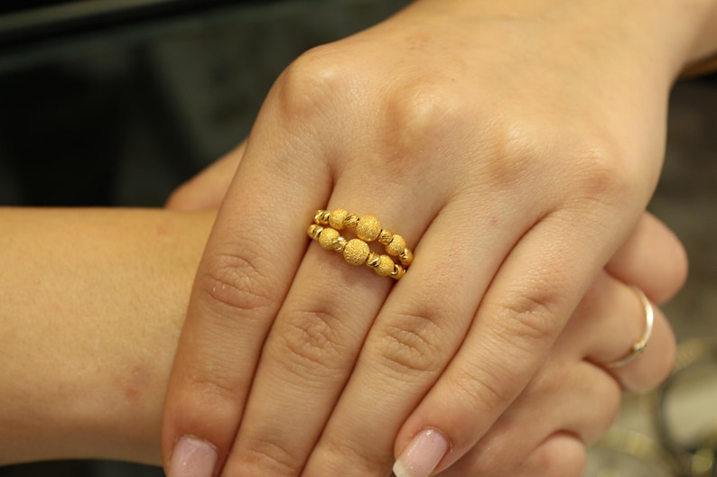 May include: A gold ring with a textured design featuring multiple small round beads. The ring is worn on a finger.