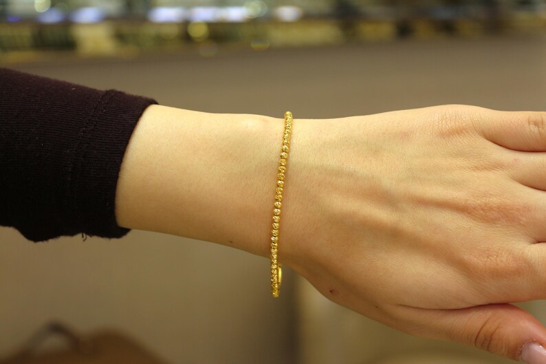 Puede incluir: Un brazalete de oro con un dise&ntilde;o de cuentas. El brazalete est&aacute; en la mu&ntilde;eca de una persona.