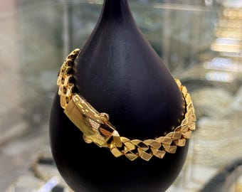 May include: A gold-colored bracelet with a curb chain design is displayed on a black teardrop-shaped stand. The bracelet features a secure clasp and a polished finish, showcasing its intricate links. The jewelry is presented against a blurred background.