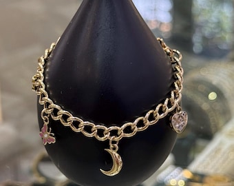Könnte beinhalten: Goldfarbenes Kettenarmband mit verschiedenen Anhängern. Zu den Anhängern gehören ein rosa-goldener Schmetterling, ein Halbmond und ein herzförmiger Anhänger mit einem klaren Stein. Das Armband wird auf einem schwarzen, tropfenförmigen Ständer präsentiert.