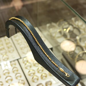 Puede incluir: Un brazalete de cadena de oro con un dise&ntilde;o texturizado que se muestra en un soporte negro. El brazalete est&aacute; en una pantalla con otras joyas.