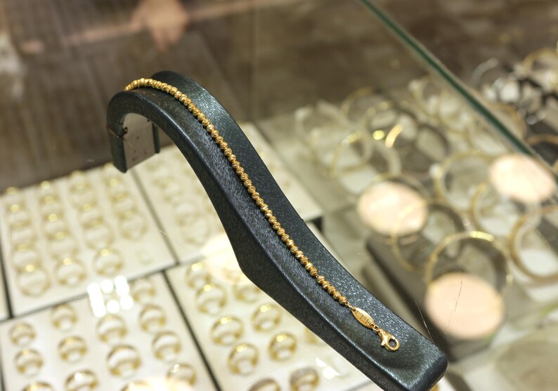 Puede incluir: Un brazalete de cadena de oro con un dise&ntilde;o texturizado que se muestra en un soporte negro. El brazalete est&aacute; en una pantalla con otras joyas.