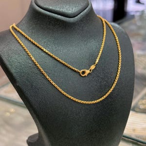 May include: A gold-toned box chain necklace featuring a polished finish and a secure lobster claw clasp. The chain consists of interconnected square links that create a smooth, rounded profile, displayed on a black neck form for clear visibility.